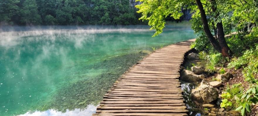 Ein schmaler Holzsteg führt über einen stillen, leicht dampfenden See mit türkisem Wasser, am Ufer frisches Grün