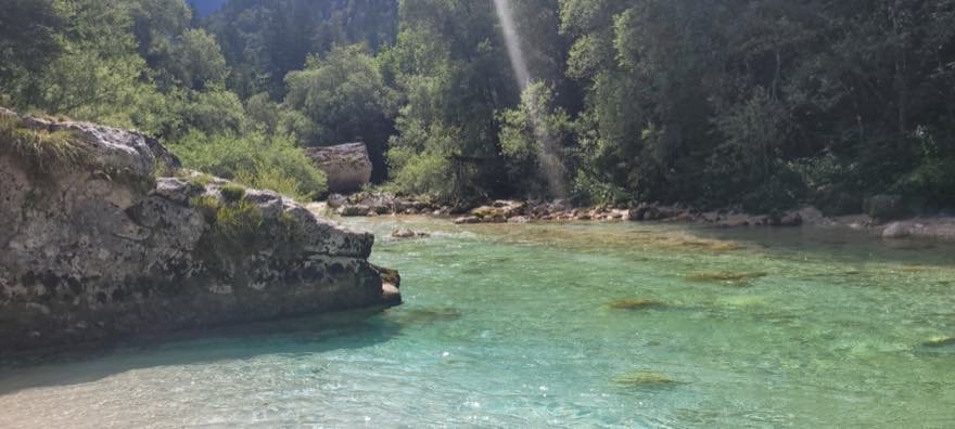 Eine ruhige Flussbucht mit klarem türkisem Wasser, davor ein flacher Felsvorsprung, im Hintergrund Wald und ein weicher Lichtstrahl